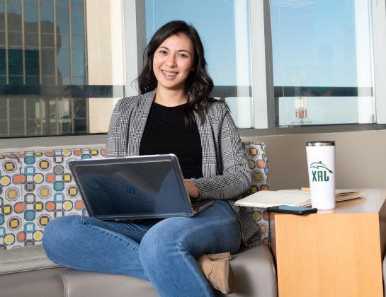 A student seating at a couch with her laptop on her hands smiles at the camera. 