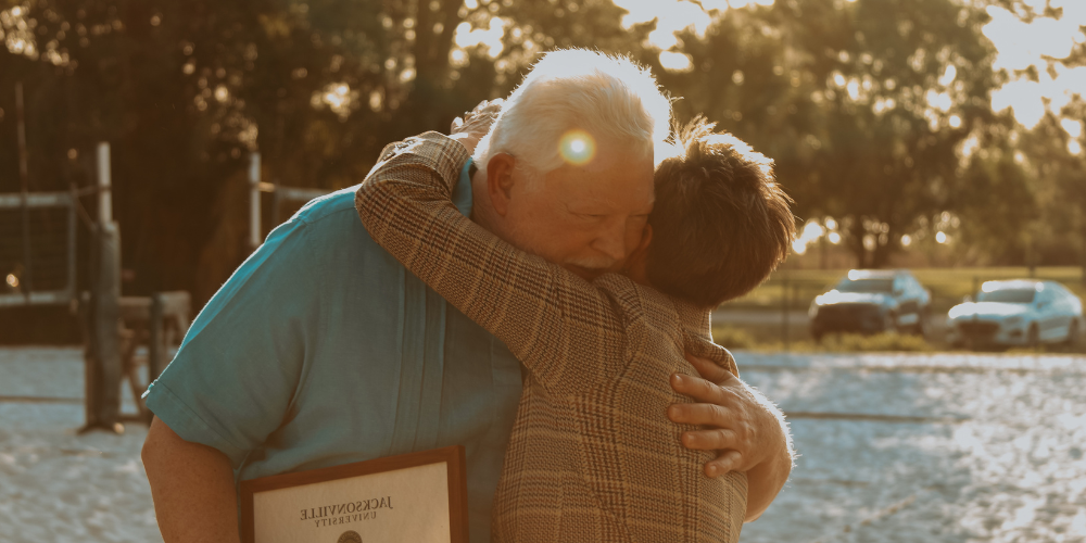 Two middle-aged adults hug at the conferring of an award against a tree-dappled, sunny, warm backdrop.
